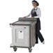 A woman in a chef hat and apron pushing a Cambro meal delivery cart with granite sand-colored trays.