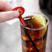 A hand holding a red WNA Comet oval stirrer in a glass of brown liquid with a lemon wedge.