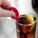 A hand holding a WNA Comet red plastic oval stirrer in a glass of brown liquid with a lemon wedge.