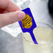 A hand holding a blue WNA Comet rectangular stirrer over a glass of lemonade.