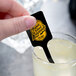 A hand holding a black rectangular stirrer over a glass of lemonade.