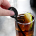 A hand holding a WNA Comet black oval drink stirrer in a glass of brown liquid with a lime wedge.