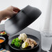 A hand using a black Cambro plate cover to keep food warm on a table.