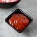 A black square Tablecraft melamine ramekin filled with ketchup on a table.