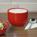 A red Fiesta jumbo china bowl filled with white liquid on a white napkin with a spoon.