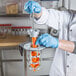 A person in a white lab coat and blue gloves using a Tellier manual upright carrot peeler.
