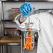 A person in a white lab coat and blue gloves using a Tellier small carrot peeler on an orange carrot.