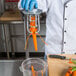 A person in a white coat and blue gloves using a Tellier manual upright small carrot peeler to peel a carrot.