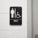 A black and white Vollrath men's restroom sign with a person and a wheelchair symbol on a white wall.