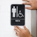 A person's hand holding a black and white sign that says "Men" with a wheelchair icon.