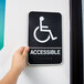 A person holding a black and white Vollrath Handicap Accessible sign with Braille.
