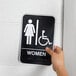 A hand holding a black and white Vollrath Women's restroom sign with Braille.