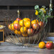 A Clipper Mill chrome plated iron wire basket filled with apples and oranges on a table.