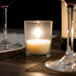 A Leola clear glass votive candle lit on a table with a bottle of wine.