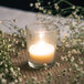 A Leola clear glass wax filled votive candle with flowers around it on a table.