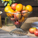 A Clipper Mill chrome plated iron wire basket filled with oranges and apples on a table.