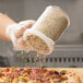 A hand using a white shaker lid to sprinkle seasoning on a pizza.