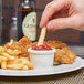 A hand dipping a french fry into a Tuxton fluted white ramekin of ketchup on a plate.