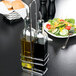 A Tablecraft metal rack holding glass bottles of oil and vinegar on a table.