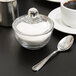 An Anchor Hocking glass sugar bowl with a lid and a spoon next to a cup of coffee.