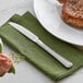 An Acopa Landsdale stainless steel dinner knife on a green napkin next to a plate of meat.