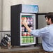 A man taking a drink from an Avantco countertop glass door refrigerator.
