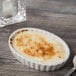 A Tuxton eggshell white oval fluted dish with brown creme brulee in it next to a spoon and glass.