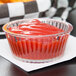 A clear fluted plastic ramekin filled with ketchup on a table.