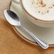 An Acopa Edgeworth stainless steel demitasse spoon on a saucer next to a cup of coffee.