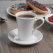 A Libbey bright white porcelain espresso saucer with a cup of coffee on it next to a plate of food.