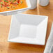 A white square Acopa porcelain saucer on a table with a bowl of soup.
