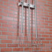 A Matfer Bourgeat utensil holder rack with three metal whisks hanging on a metal pole against a brick wall.