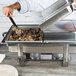 A hand using a Vollrath hinged dome cover to prepare food in a large stainless steel pan on a table.