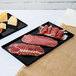 Two black Tablecraft cast aluminum rectangular cooling platters of meat and cheese on a counter.