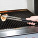 A person using Vollrath Jacob's Pride tongs with a black handle to cook food on a grill.