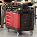 A man pushing a black Rubbermaid TradeMaster tool cart with drawers and a cabinet.