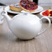 A white Schonwald teapot with a lid on a table next to a bowl of fruit.