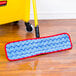 A red and blue Rubbermaid HYGEN wet mop pad attached to a mop handle on a wood floor.