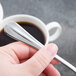 A hand holding a Libbey stainless steel teaspoon over a cup of coffee.
