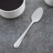 A Libbey stainless steel teaspoon next to a cup of coffee on a table.