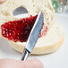A hand using a Libbey stainless steel bread and butter knife to spread jam on a slice of bread.