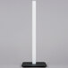 A white pole on a black square stand holds a tall white candle.