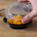 A person's finger touching a Fabri-Kal plastic container filled with oranges and covered with a vented lid.