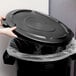 A person putting a black plastic lid on a Rubbermaid commercial trash can.