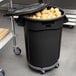 A Rubbermaid black round trash can with a lid and dolly filled with potatoes.