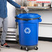 A man standing next to a blue Rubbermaid recycling bin with a recycling lid.