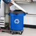 A person putting a plastic container into a blue Rubbermaid recycling bin with a white lid.