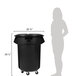 A woman standing next to a black Rubbermaid BRUTE trash can with wheels.