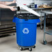 A woman using a white lid to put recyclables in a blue Rubbermaid BRUTE recycling can.