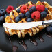 A waffle with berries and chocolate syrup on a black plate with a Chef & Sommelier stainless steel dessert knife.